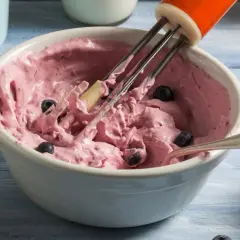 Różowy mus jagodowy do tortu, przygotowywany mikserem, z widocznymi całymi jagodami.