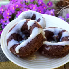 Trzy czekoladowe donuty brownie polane białym lukrem, na talerzu wśród fioletowych kwiatów.