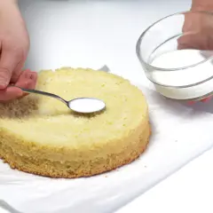 Ręce dodają łyżeczką płynny poncz kawowy do biszkoptu, przygotowując go do dalszego dekorowania.