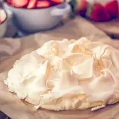 Beza na papierze do pieczenia, z widocznymi naczyniami z truskawkami w tle. Zastanawiasz się, dlaczego beza pęka?