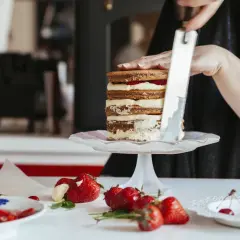 Osoba dekoruje tort z masła, dodając krem. Na stole świeże truskawki i wiśnie.