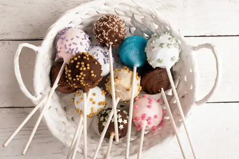 Kolorowe cake pops kokosowe w białym koszyku, udekorowane posypkami i kuleczkami.