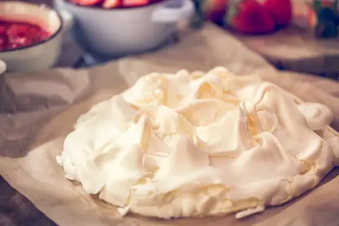 Beza na papierze do pieczenia, z widocznymi naczyniami z truskawkami w tle. Zastanawiasz się, dlaczego beza pęka?