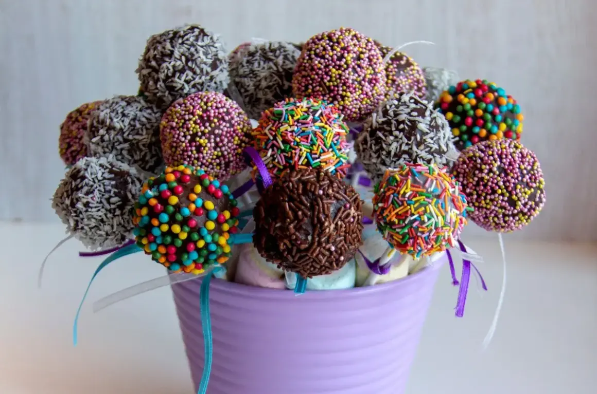 Kolorowe cake pops z jasnego biszkoptu, udekorowane posypkami i wiórkami kokosowymi, w fioletowym wazonie.