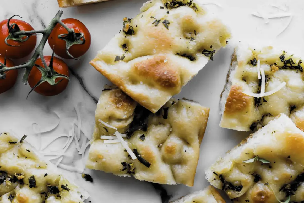 Pyszna focaccia z ziołami i parmezanem, idealna do przygotowania według prostego przepisu. Obok gałązka pomidorków koktajlowych.