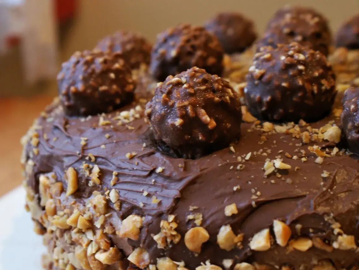 Pyszny tort oblany czekoladą, ozdobiony całymi pralinami i posypany orzechami. Wygląda jak idealny krem Ferrero Rocher.