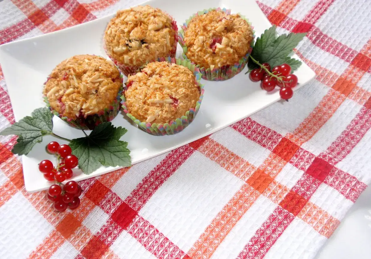 Cztery muffinki z czerwoną porzeczką, posypane płatkami owsianymi, na białej tacy z gałązkami czerwonej porzeczki.