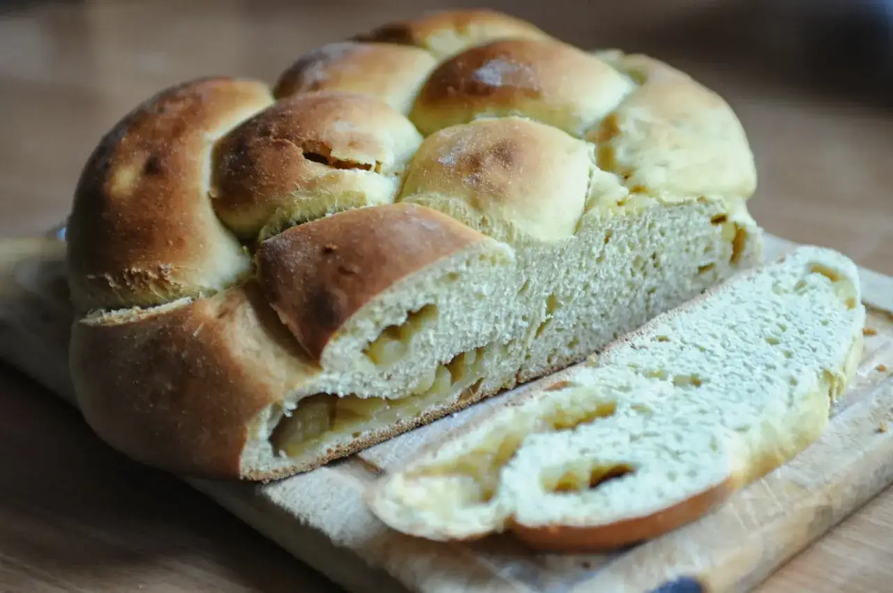Puszysta chałka z jabłkami, zapleciona i lekko zarumieniona, z widocznymi kawałkami jabłek w środku. Idealna na śniadanie.