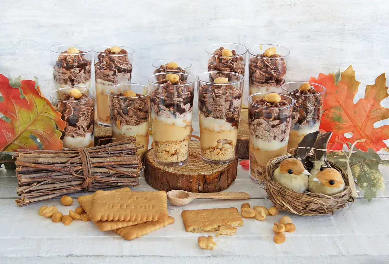 Deserki w pucharkach z warstwami kremu, chrupek i polewy, przypominające smakiem salted caramel. Obok ciasteczka i orzeszki.
