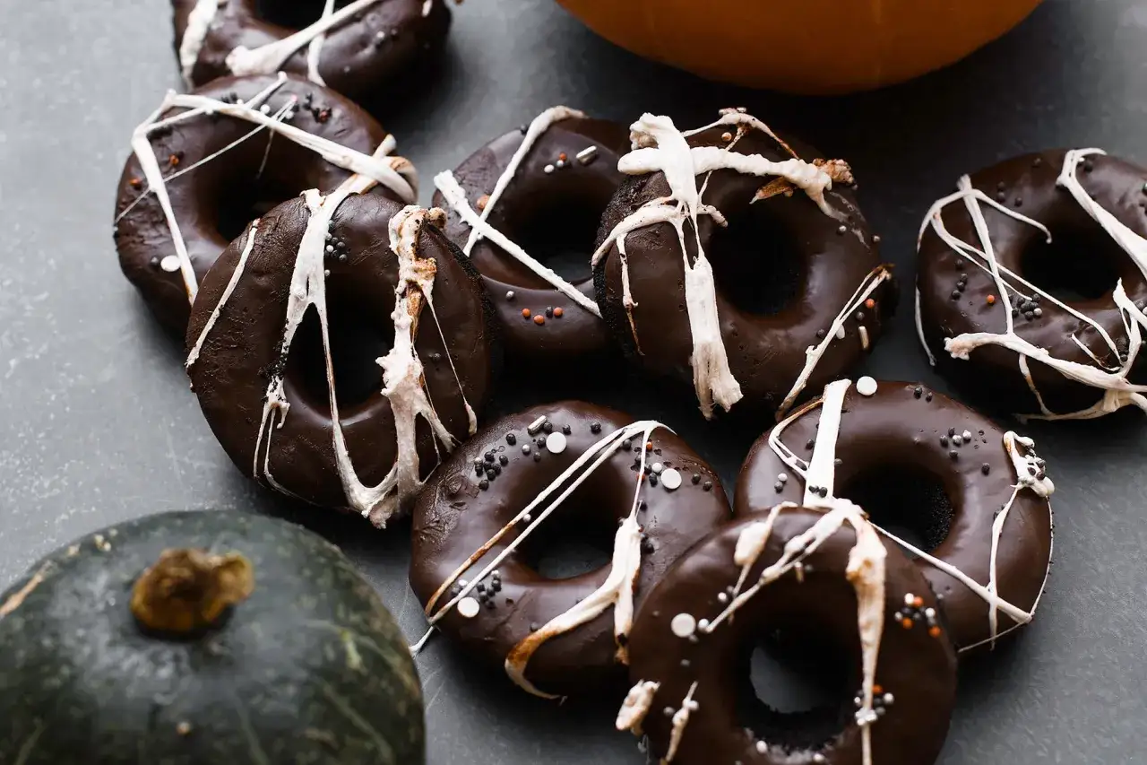 Ciemne, czekoladowe donuty brownie ozdobione białym lukrem i posypką, obok małe dynie.