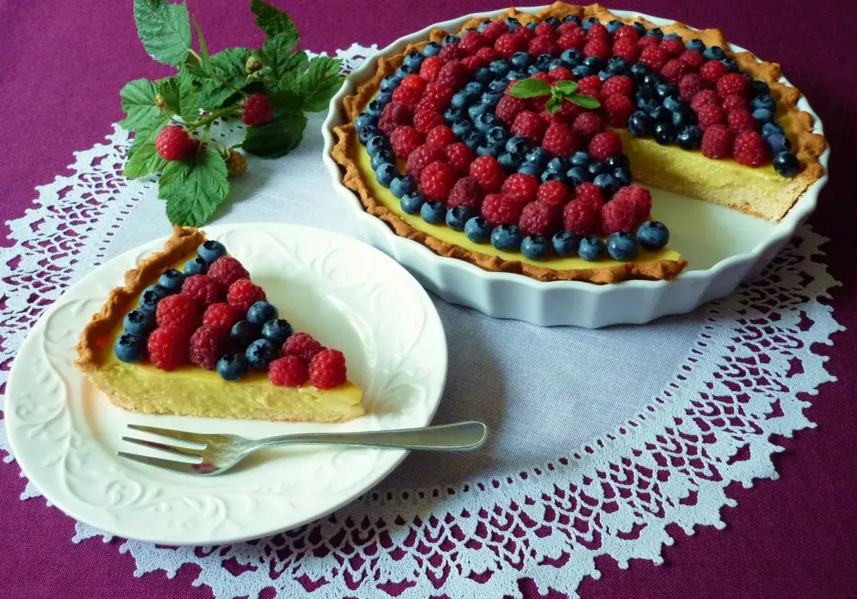 Pyszna tarta z kremem patissiere, ozdobiona malinami i bor&oacute;wkami. Kawałek na talerzyku czeka na spr&oacute;bowanie.