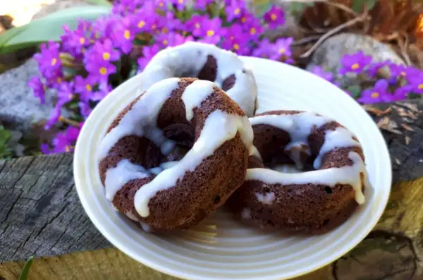 Trzy czekoladowe donuty brownie polane białym lukrem, na talerzu wśród fioletowych kwiatów.