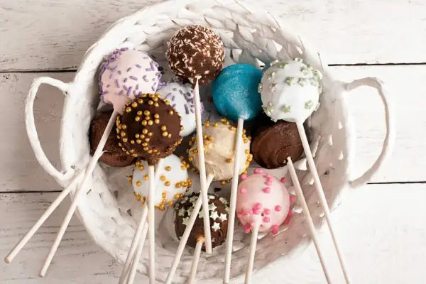 Kolorowe cake pops kokosowe w białym koszyku, udekorowane posypkami i kuleczkami.