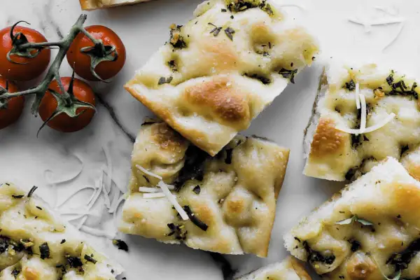 Pyszna focaccia z ziołami i parmezanem, idealna do przygotowania według prostego przepisu. Obok gałązka pomidorków koktajlowych.