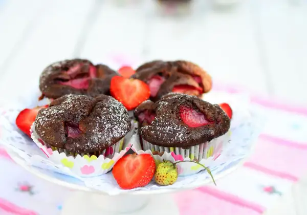 Pyszne muffiny czekoladowe z truskawkami, posypane cukrem pudrem, prezentują się apetycznie na talerzyku.
