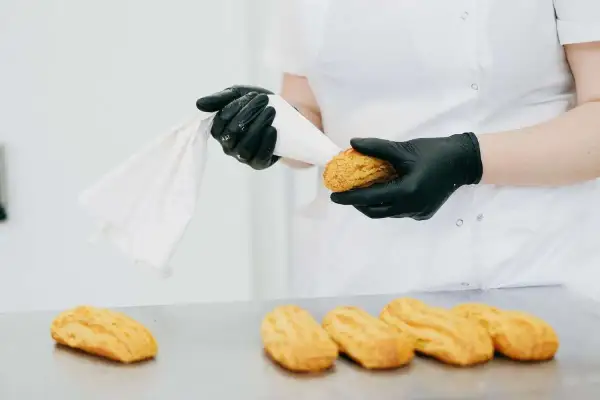 Ręce w czarnych rękawiczkach przygotowują ptysie do nadzienia kremem z żółtek. Na stole leżą gotowe ciastka.
